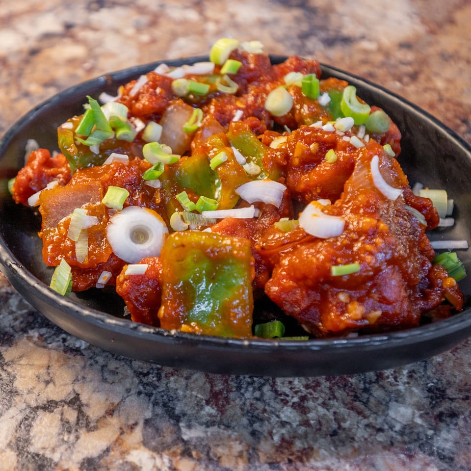 Chili chicken with green bell peppers and scallions in a black pan on a marbled countertop.
