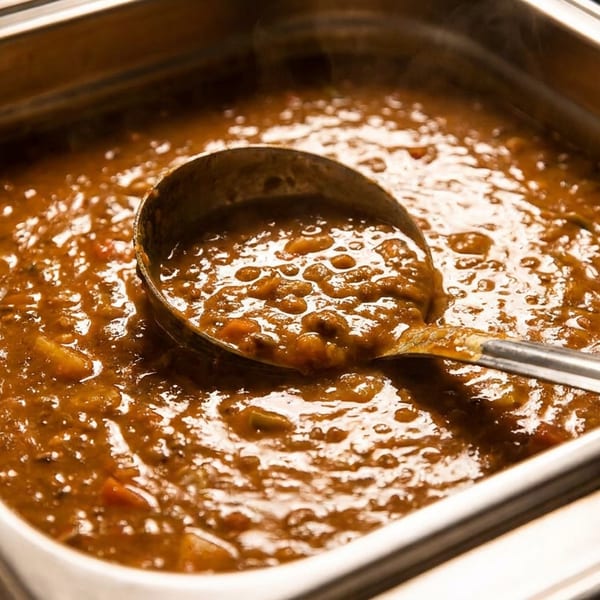 Thick brown lentil stew in a metal buffet pan with a ladle, steaming hot.