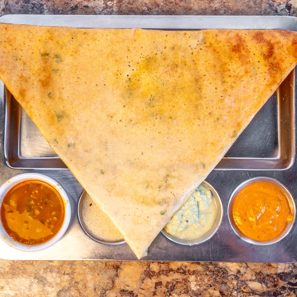Golden-brown triangular dosa served with three colorful dipping sauces on a metal tray.
