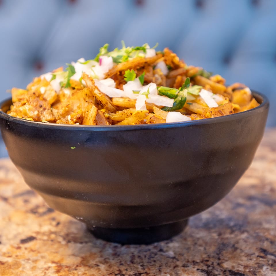 Black bowl of seasoned potato pieces, garnished with chopped onions, cilantro, and green chilies.